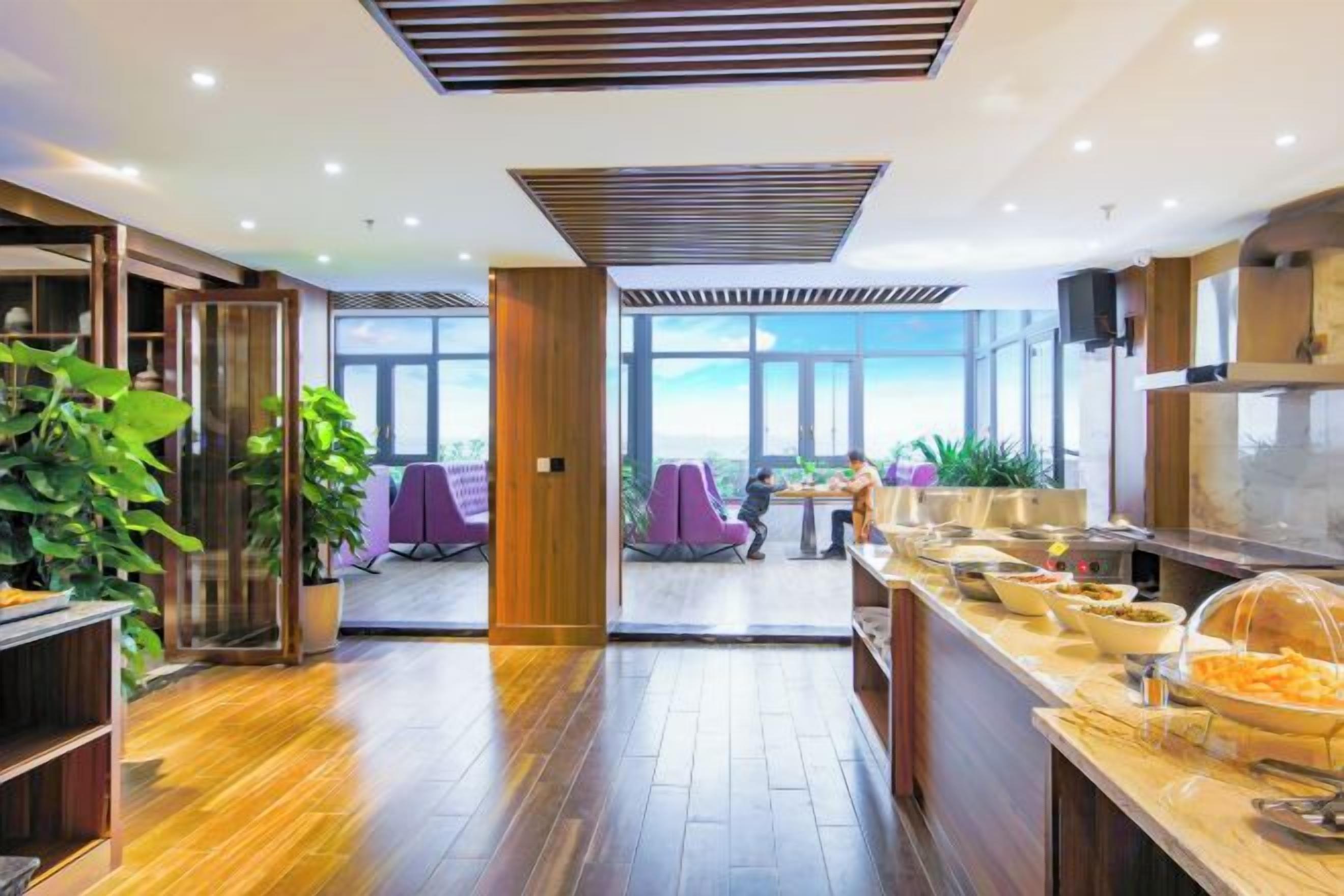 A modern hotel lobby with a wooden floor, a buffet area with various food items, and a view of the cityscape through large windows.