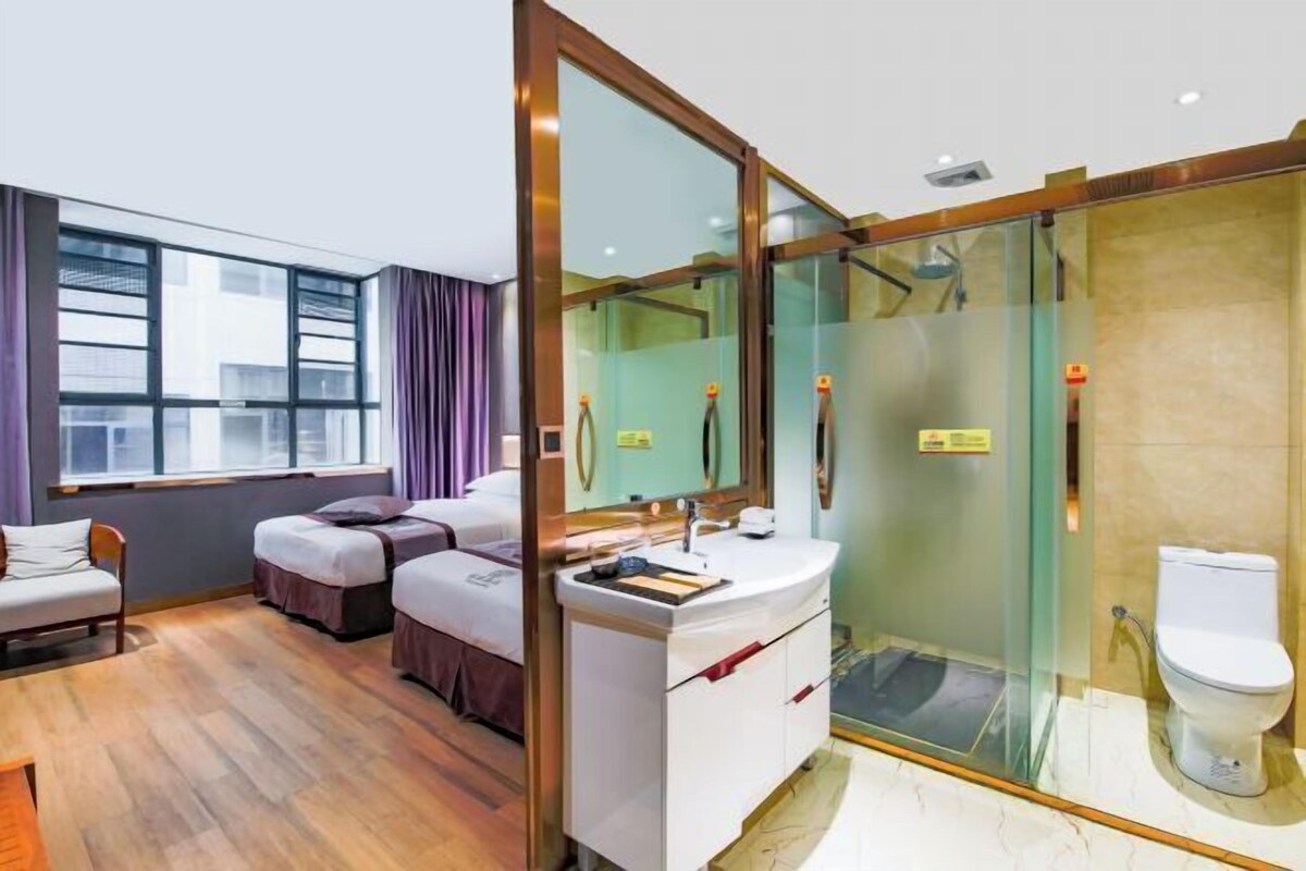A hotel room with a large window, a shower with glass doors, a white sink, and a bed with two pillows.