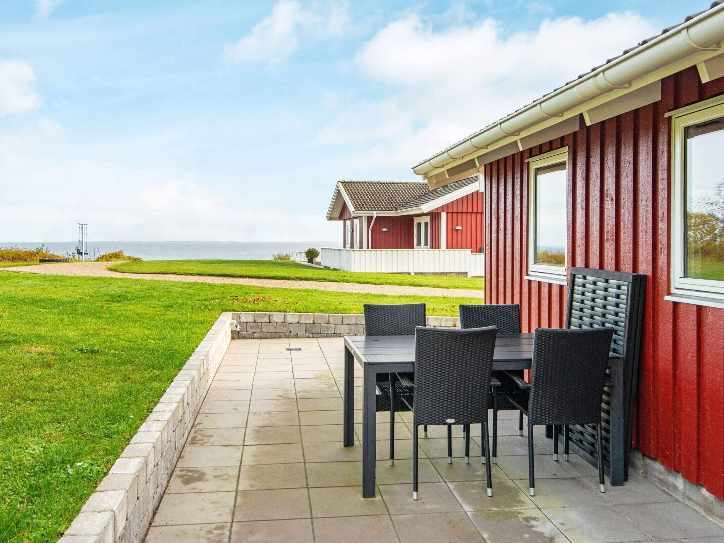 A red building with a patio area featuring a table and chairs, a view of the sea, and a grassy landscape.