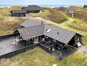 Een modern houten huis met een ruim terras, omgeven door duinen en de zee.
