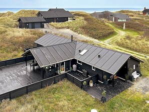 Ett modernt trähus med en stor terrass, omgivet av sanddyner och havet.