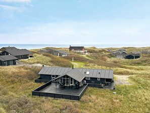 Ett byggnadskomplex vid kusten med flera olika byggnader, inklusive ett huvudhus med en stor terrass och en mindre byggnad med ett sluttande tak.