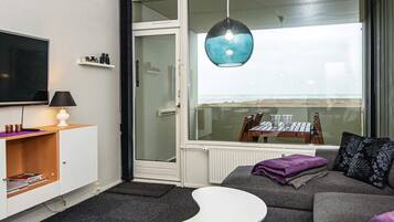 A modern living room with a flat-screen TV, a white coffee table, a grey sofa, and a large window with a view of the beach.