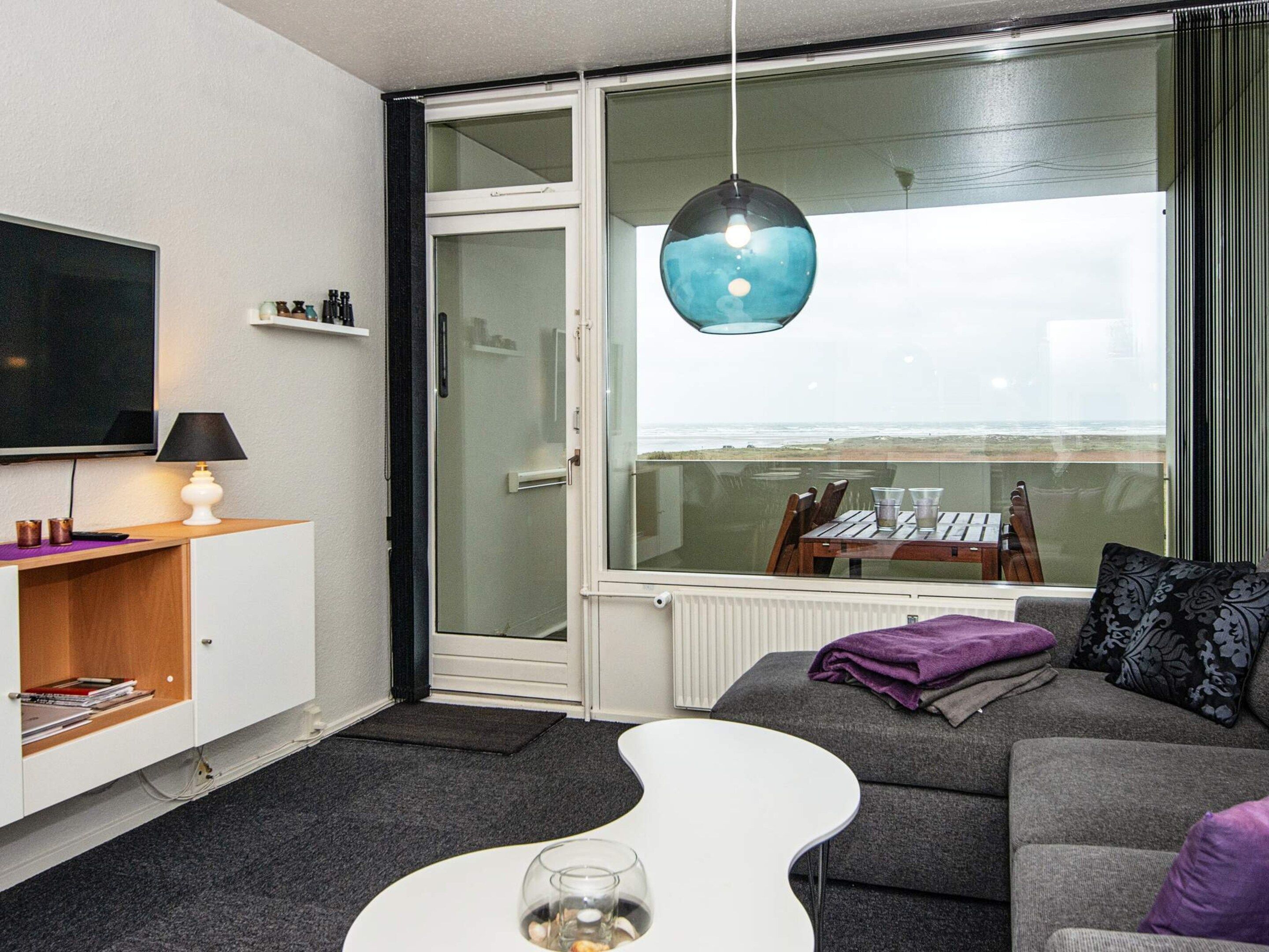 A modern living room with a large TV, a glass door leading to a balcony, and a view of the sea.