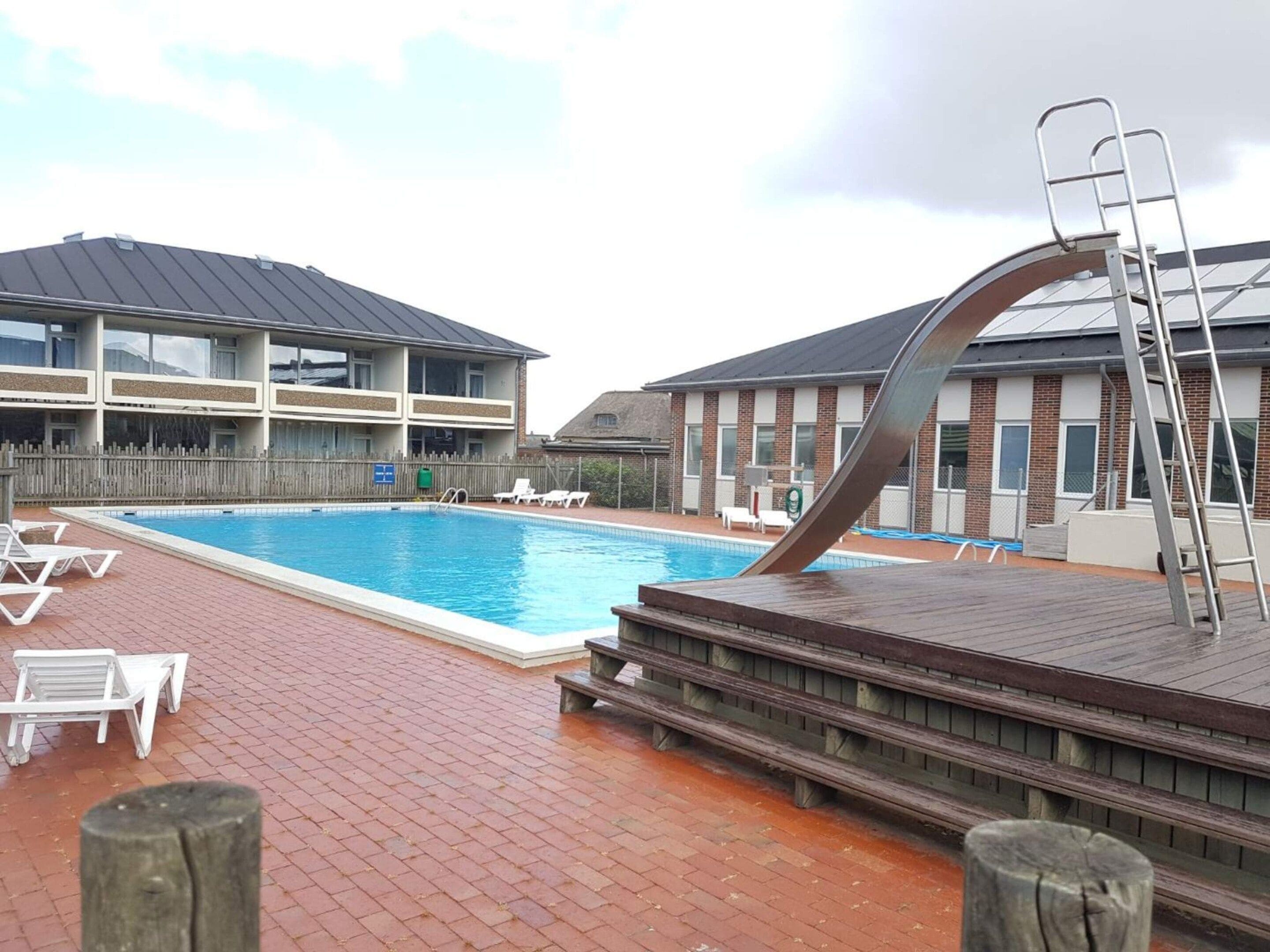 Ein Schwimmbecken mit Holzterrasse und Liegestühlen, umgeben von Gebäuden mit Backsteinfassaden und großen Fenstern.