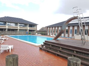 Ein Schwimmbecken mit Holzterrasse und Liegestühlen, umgeben von Gebäuden mit Backsteinfassaden und großen Fenstern.