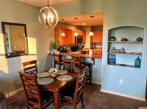 A dining area with a wooden table and chairs, a chandelier, and a kitchen with wooden cabinets and a blue accent wall.