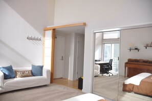 Un dormitorio moderno con una cama grande, un sofá blanco con cojines azules, un escritorio de madera y un ventanal de vidrio que da a otra habitación.