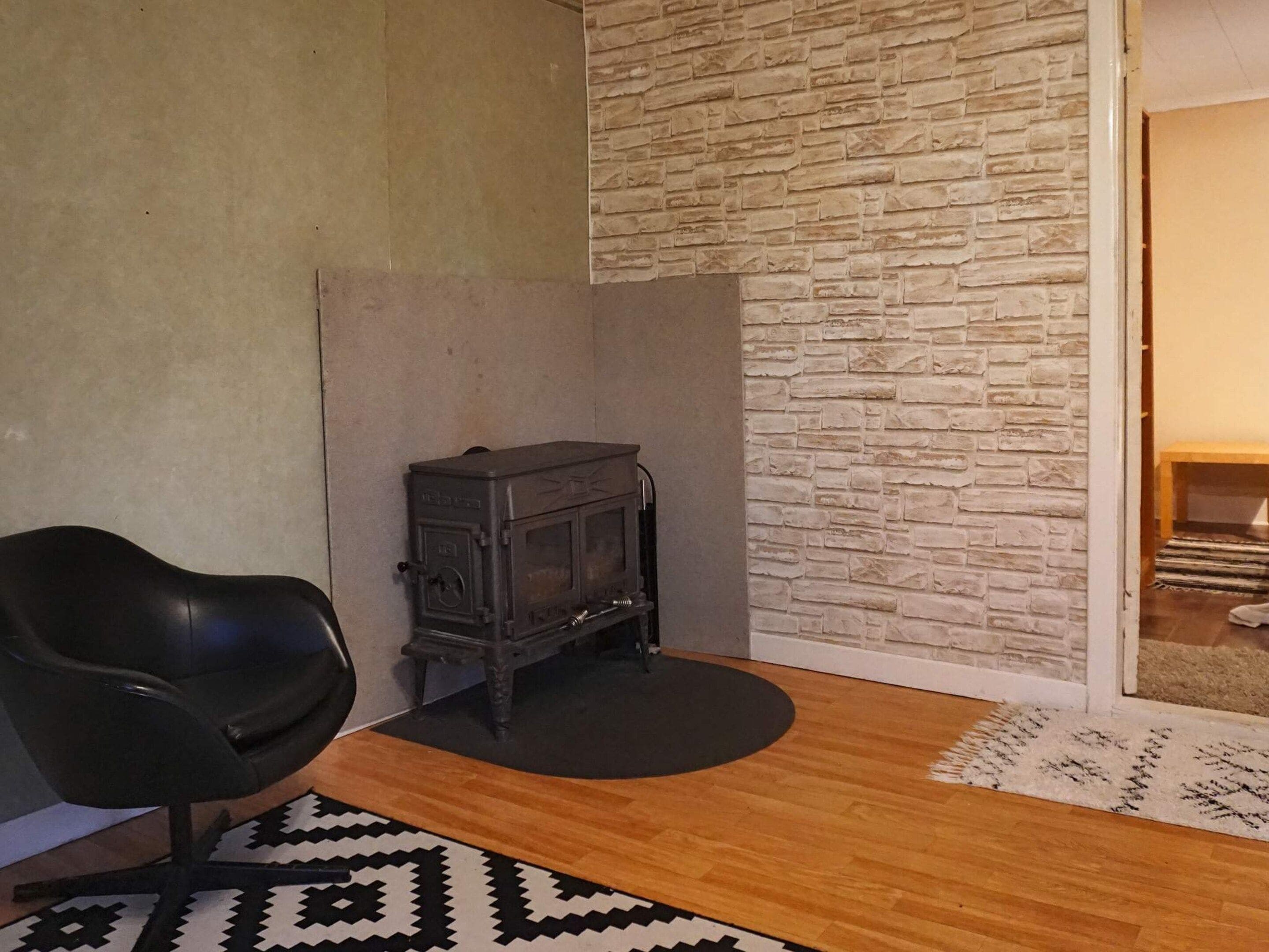 Una habitación con un sillón de cuero negro, un pequeño calentador negro, un tapete estampado y una pared con textura de piedra.