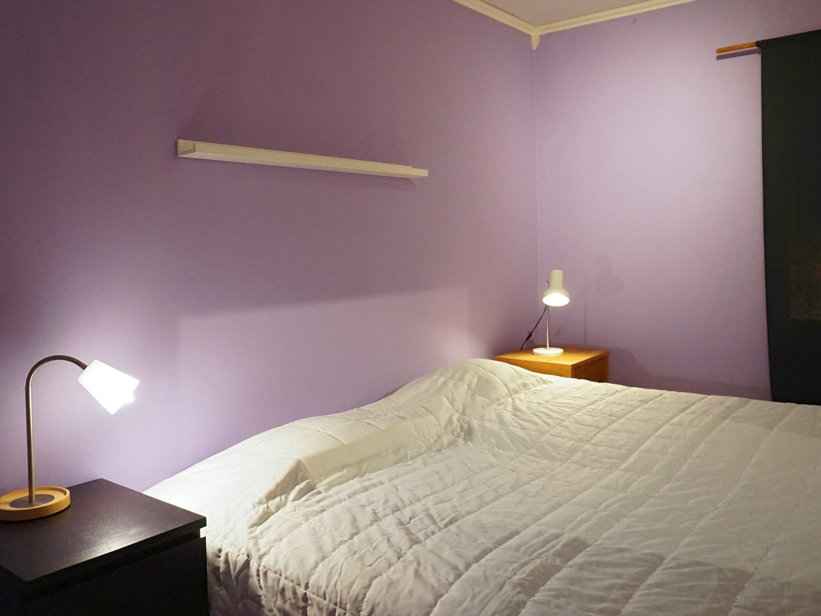 A bedroom with a bed, a nightstand with a lamp, and a wall-mounted shelf.