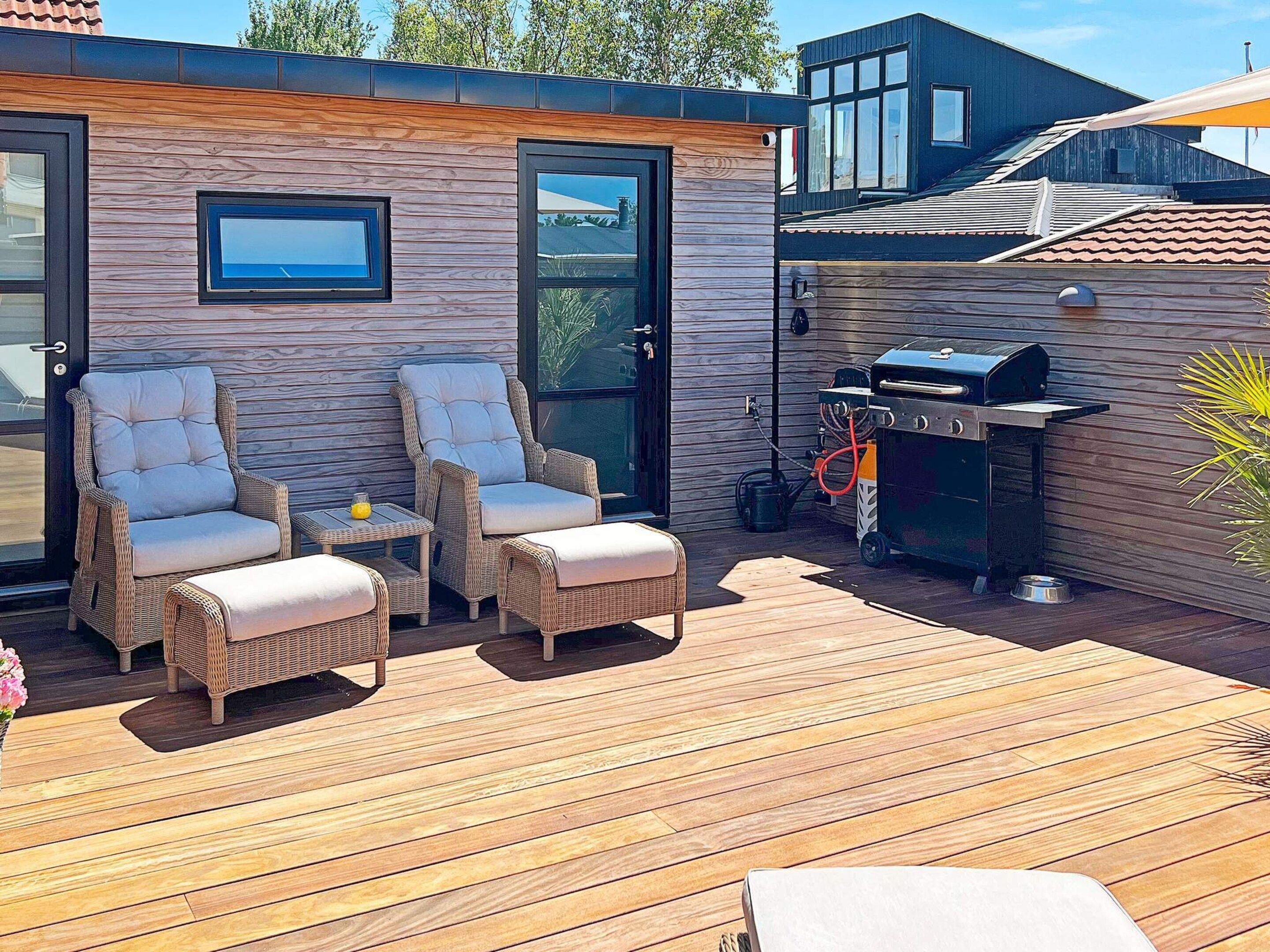 An outdoor deck with wicker furniture, a grill, and a wooden wall with a window.