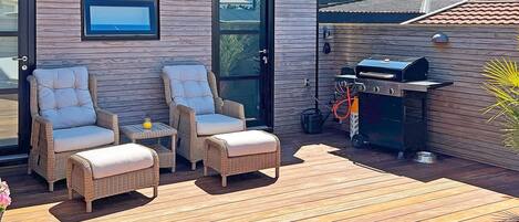 An outdoor deck with wicker furniture, a grill, and a wooden wall with a window.