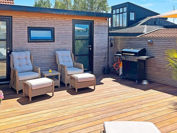 Une terrasse extérieure avec des meubles en osier, un barbecue et un mur en bois doté d’une fenêtre.