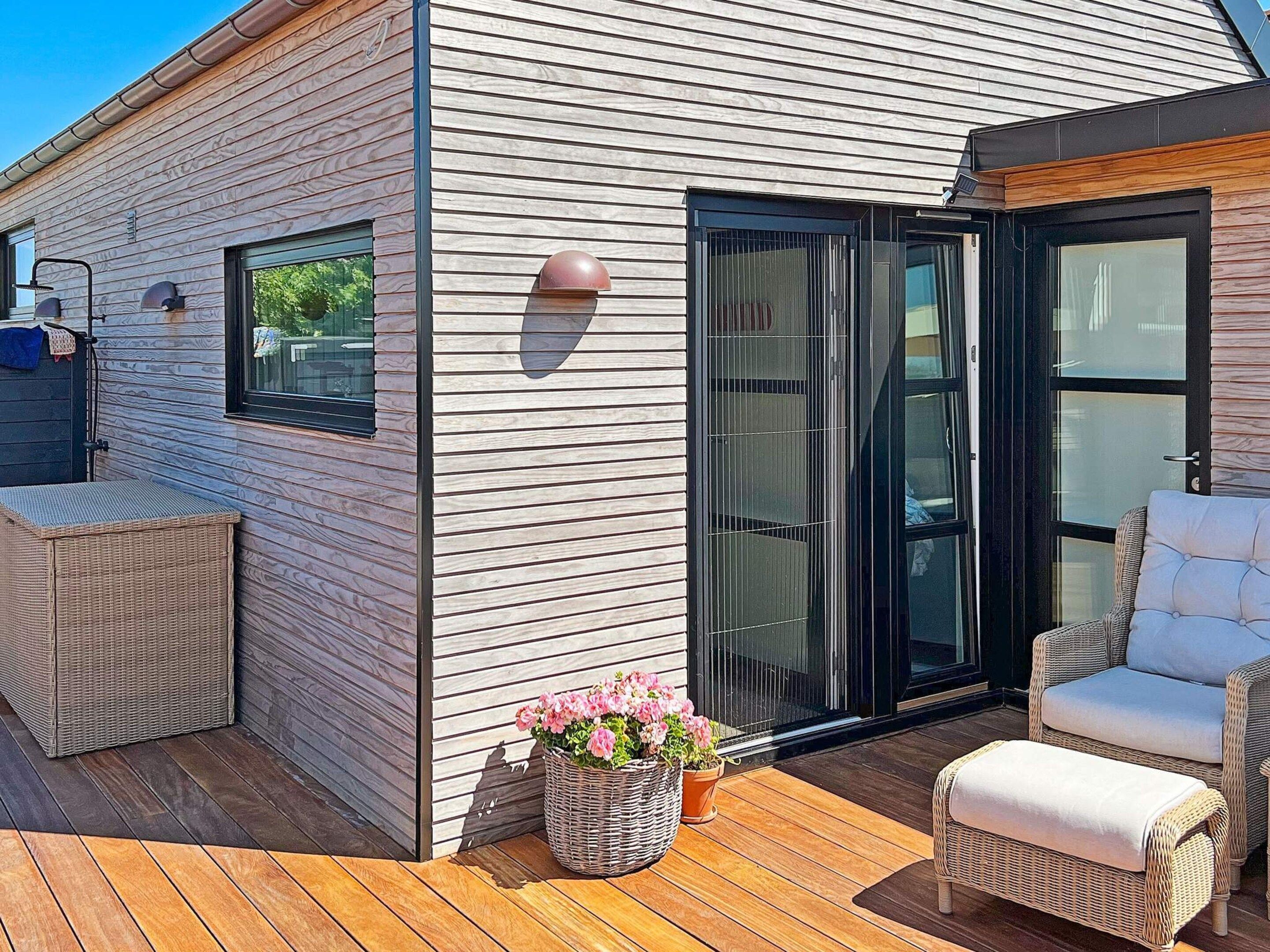 Een modern exterieur met een houten gevel, een schuifglazen deur, tuinmeubels van riet en potplanten.