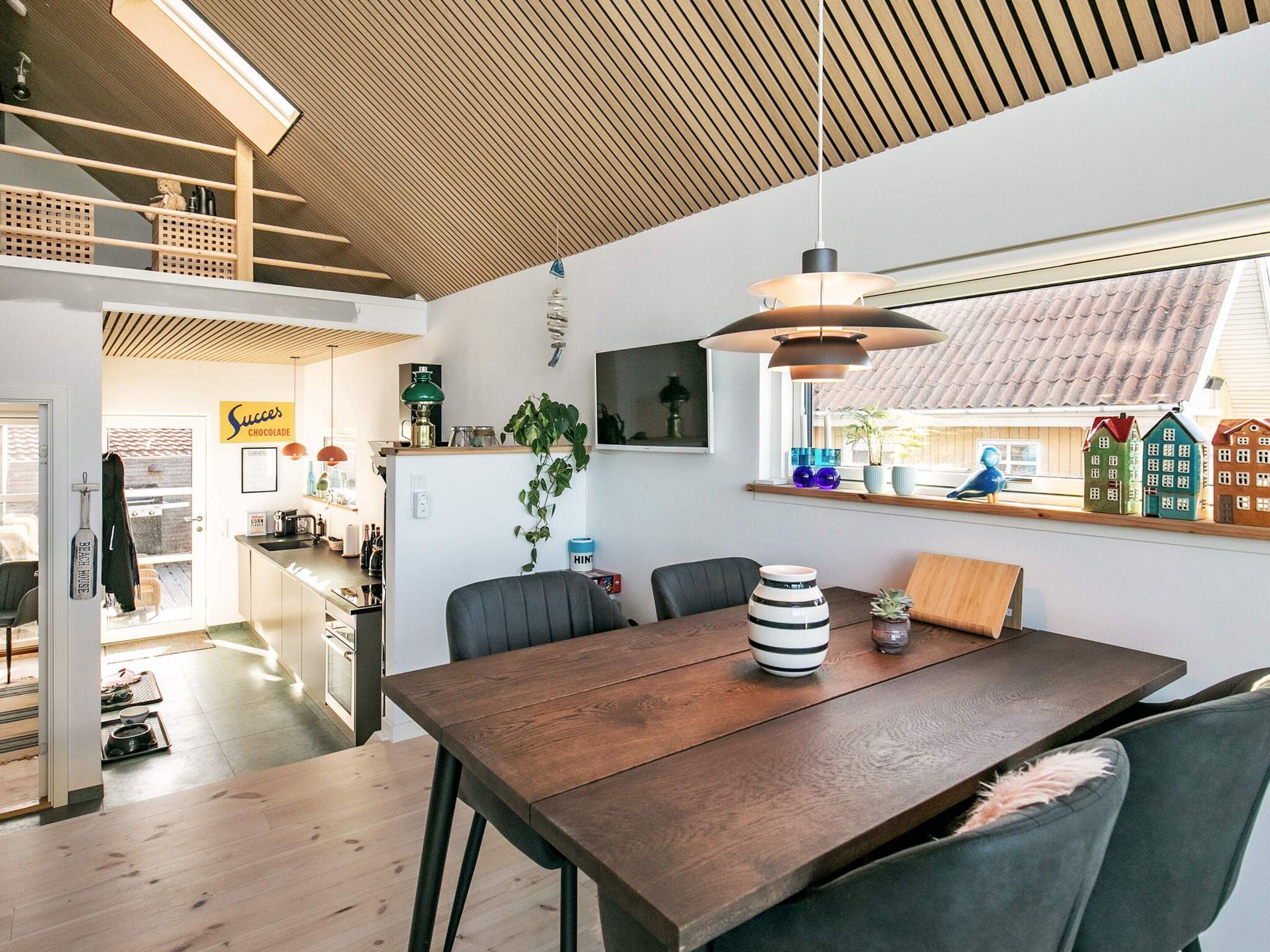 A modern kitchen with a wooden ceiling, a dining area with a wooden table, and a view of houses through a window.