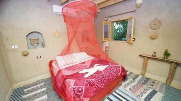 A room with a bed covered in a red canopy, a small window, a wall-mounted shelf with decorative items, and a patterned rug.
