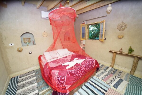 Una habitación con una cama cubierta por un dosel rojo, una ventana pequeña, un estante en la pared con objetos decorativos y una alfombra estampada.