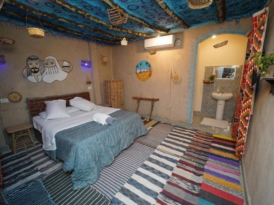 Una habitación con cama, un taburete pequeño, un estante en la pared, una cesta colgante y un baño con lavamanos y espejo.