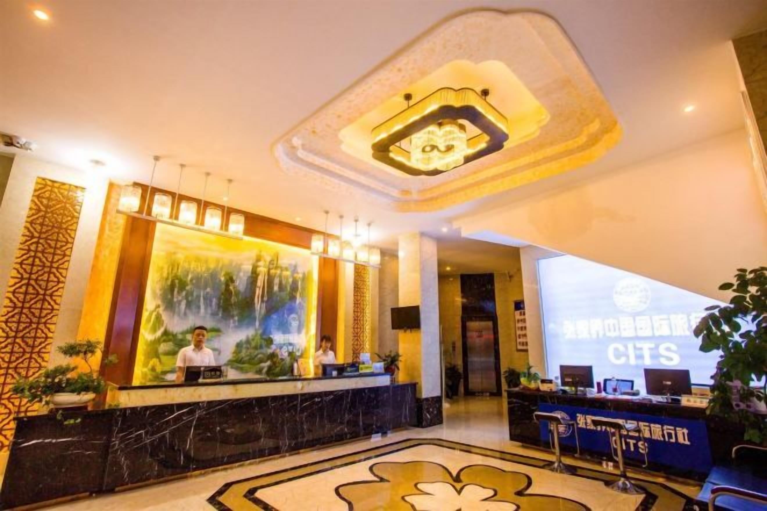 A hotel lobby with a reception desk, a large digital screen displaying the hotel's name, and a modern ceiling design.