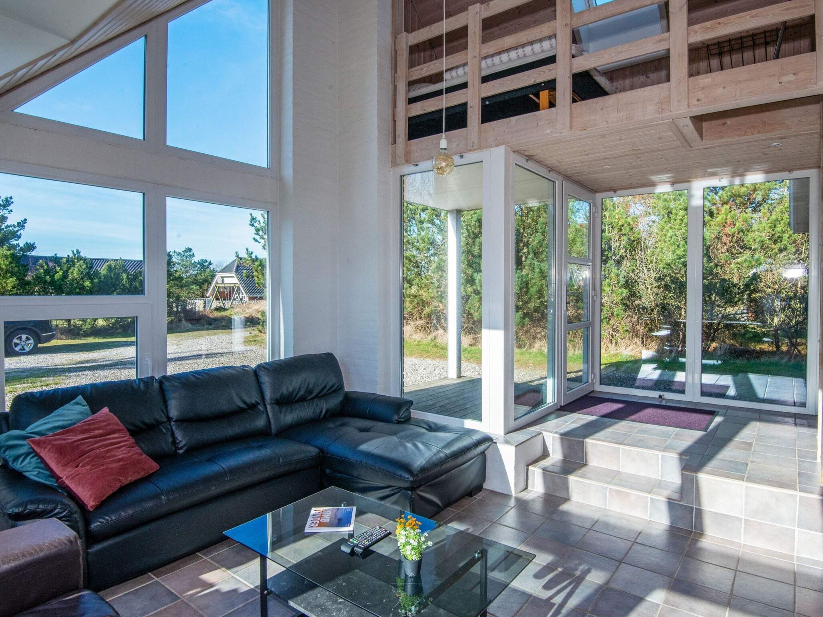 A modern living room with a black leather sofa, a glass coffee table, and large windows offering a view of the outdoors.