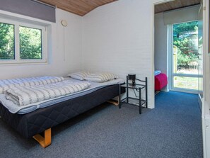 A single bed with a quilted mattress, a small table with a lamp, and a view of greenery through a door. - 4 Star Holiday Home in Romo (Rømø)