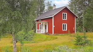 A red wooden house with a white porch and a chimney, surrounded by lush greenery and a field.