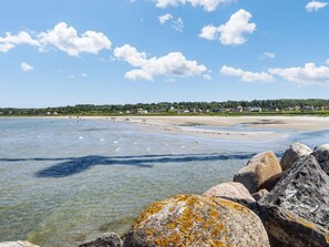 A coastal beach with clear water, rocks, and houses in the background.