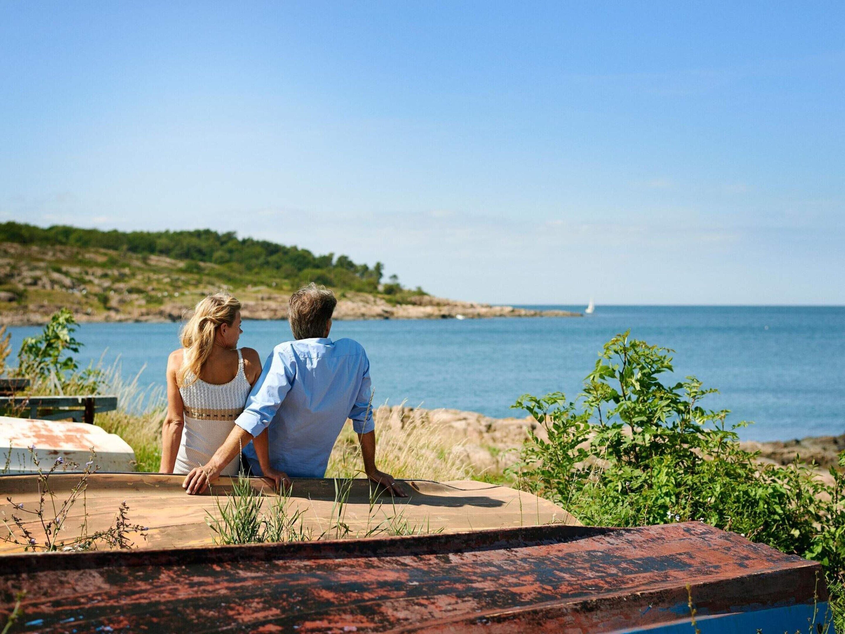 Una pareja sentada en un banco, con vista al mar y una barandilla metálica rústica al frente.