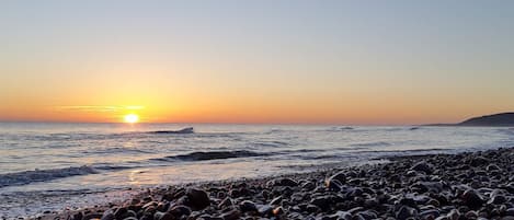 En strand med småsten och solen som går ner över vattnet.