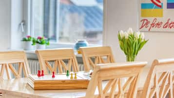 Una mesa de comedor de madera con piezas de ajedrez, un jarrón de flores y un ventanal con vistas a edificios.