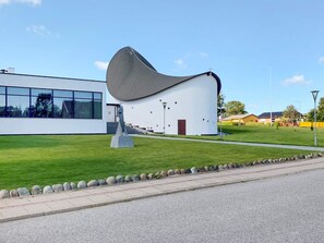 A modern building with a unique curved roof, a clear glass facade, and a well-maintained lawn. - 6 Person Holiday Home in Strandby (Strandby)
