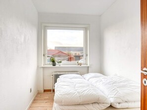 Um quarto pequeno e luminoso com uma cama, um radiador e uma janela com vista para uma área residencial.