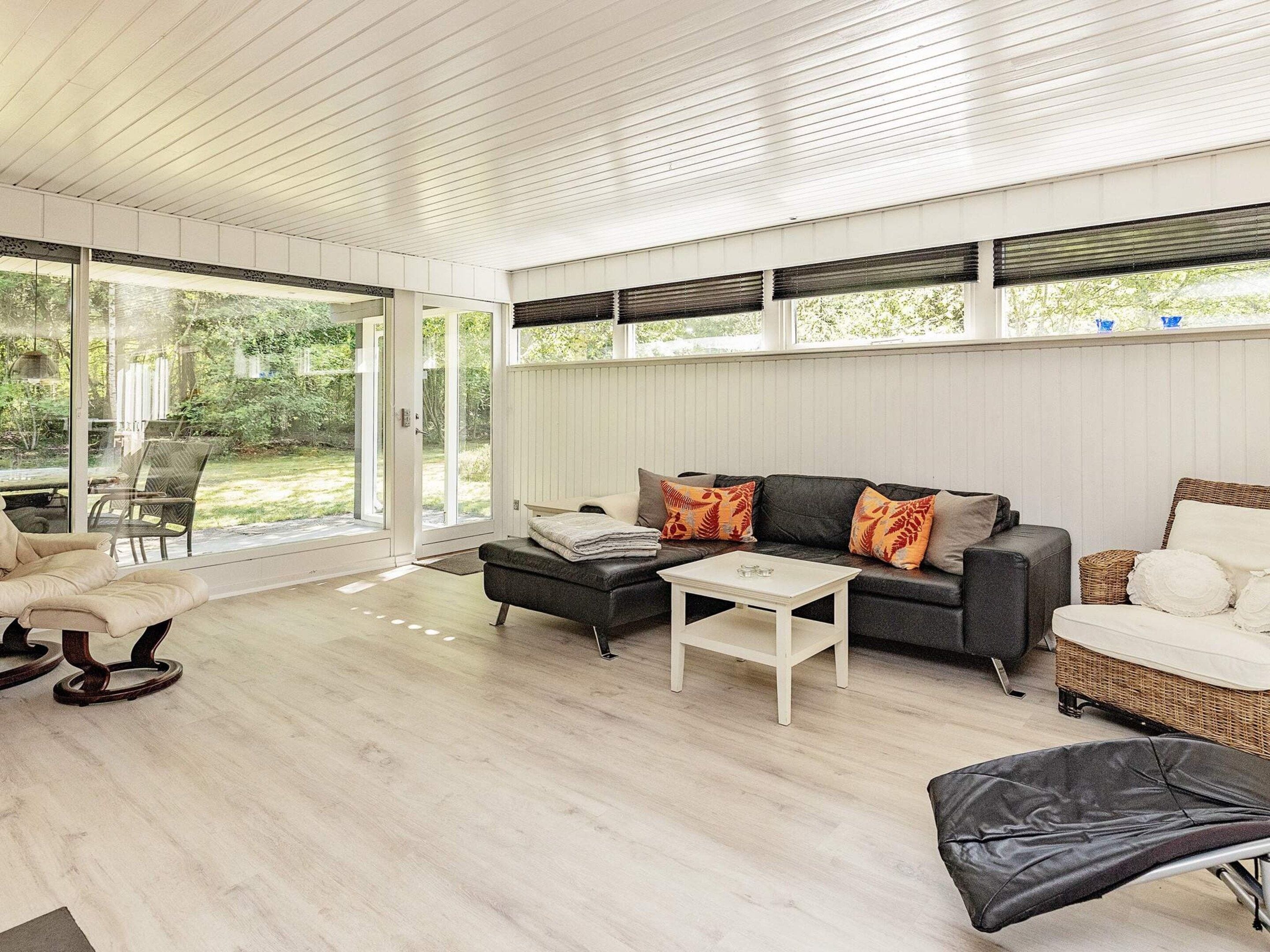 Una sala de estar moderna con sofá, reposapiés y mesa de centro, ventanas amplias con persianas, y vista a áreas verdes.