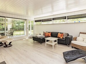 A modern living room with a sofa, ottoman, and coffee table, large windows with blinds, and a view of greenery outside. - 6 Person Holiday Home in Hojslev (Hojslev)