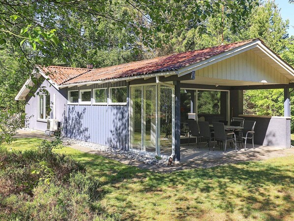 Een overdekt terras met een tafel en stoelen, een klein gebouw met een raam, en een tuin met gras en struiken.