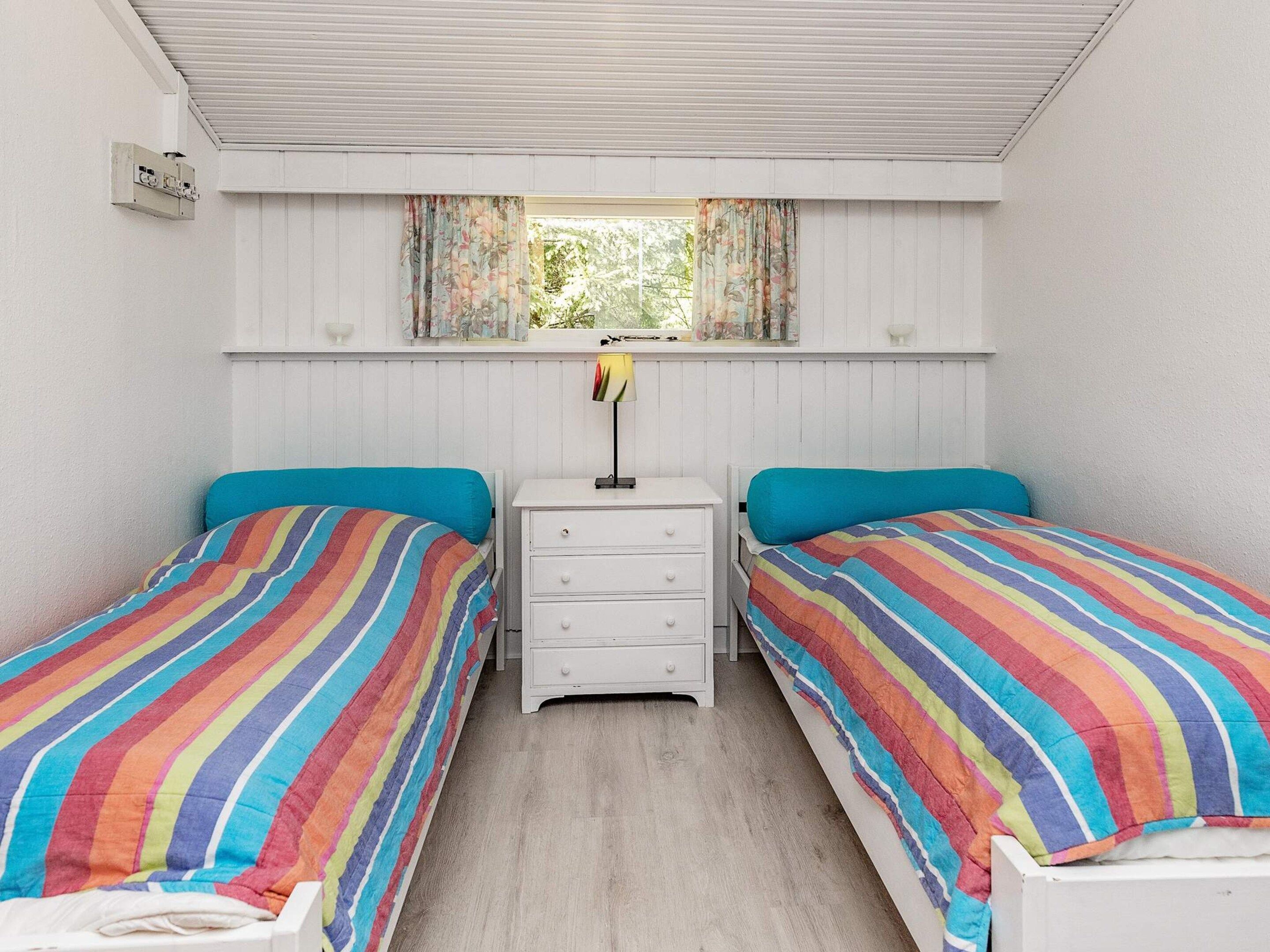Una habitación con dos camas individuales, una mesita de noche pequeña con cajones, una ventana con cortinas y un respiradero en el techo.