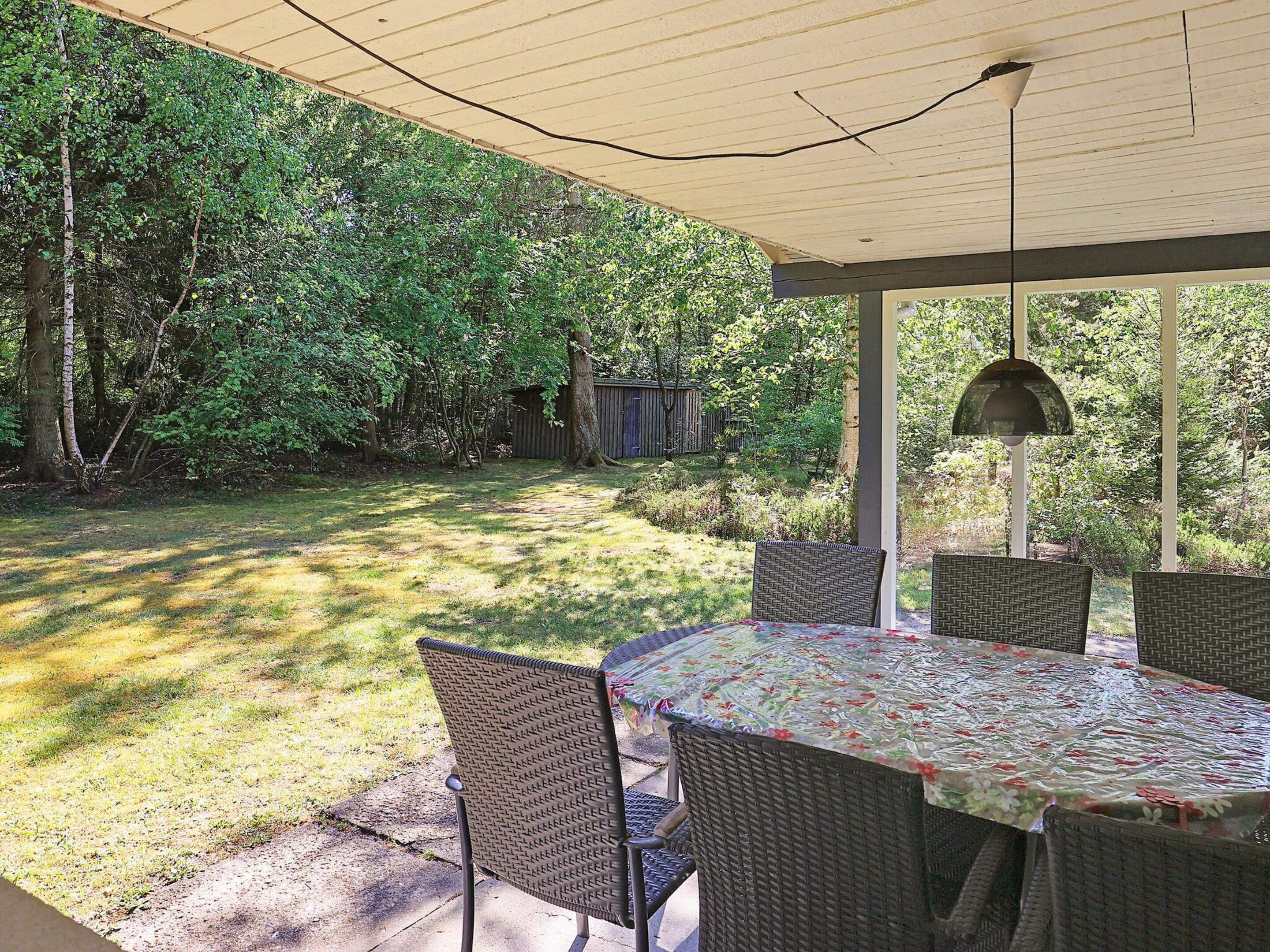 A covered patio with a table and chairs, surrounded by trees and a wooden structure in the background.