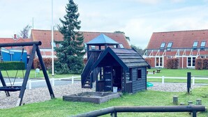 Une aire de jeux avec des balançoires et une petite maisonnette en bois dotée d’un toit bleu.