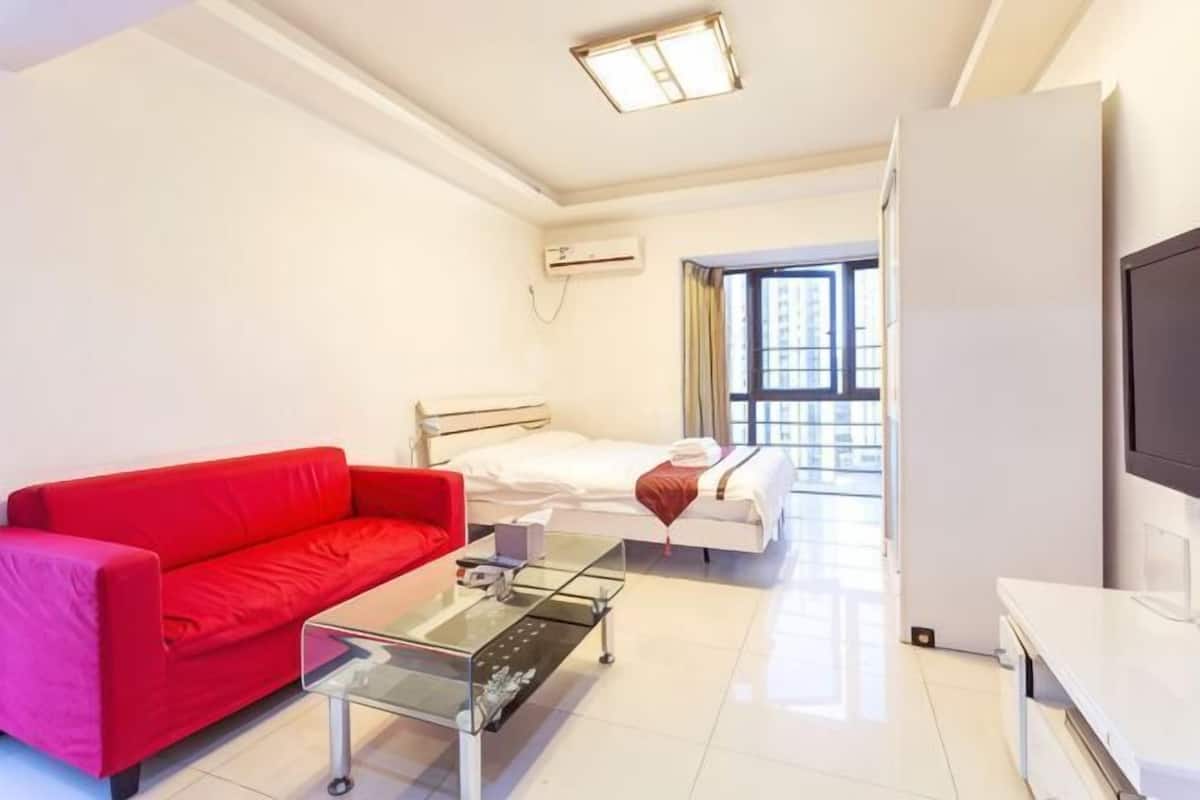 Una habitación de hotel con un sofá rojo, una mesa de centro de vidrio, una cama con ropa de cama blanca, un televisor y un ventanal grande.