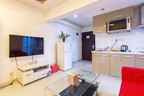 A modern living room with a flat-screen TV, a glass coffee table, a red couch, and a kitchen area with cabinets and a microwave.