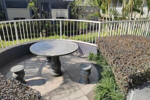 Una mesa redonda al aire libre con cuatro taburetes, rodeada por una valla de metal blanco, sobre una zona pavimentada con guijarros y plantas.