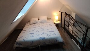 Een kleine kamer met een schuin dak, een bed, een nachtkastje, een lamp en een houten vloer.