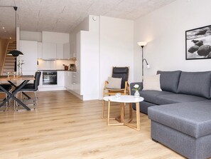 En moderne stue med en grå sofa, et træspisebord og et køkken med hvide skabe.