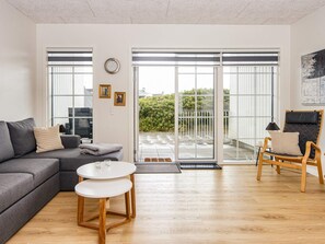 Una sala de estar moderna con un sofá gris, una mesa redonda y una silla de madera. Hay ventanas amplias con persianas, una puerta que da al exterior y una pequeña mesita con una lámpara.
