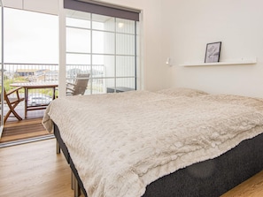A bedroom with a large bed, a chair, a small table, a balcony with a view, and a picture on the wall.