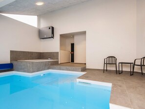 En moderne indendørs swimmingpool med et klart, blåt vandelement, et siddeområde med stole og en dør, der fører til et andet rum.