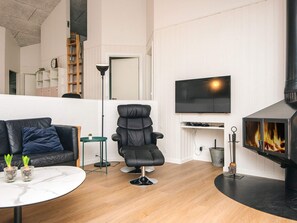 En moderne stue med en lædersofa, en sort lænestol, et rundt sofabord med planter, et fladskærms-tv monteret på væggen og en brændeovn.