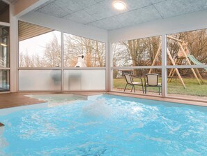En indendørs swimmingpool med glasvægge, et trædeck og udsigt til en legeplads udenfor.