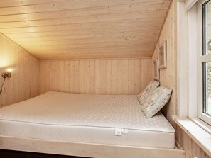 Un llit individual en una habitació de fusta amb sostre inclinat, una làmpada penjada a la paret i una finestra amb vista a vegetació.