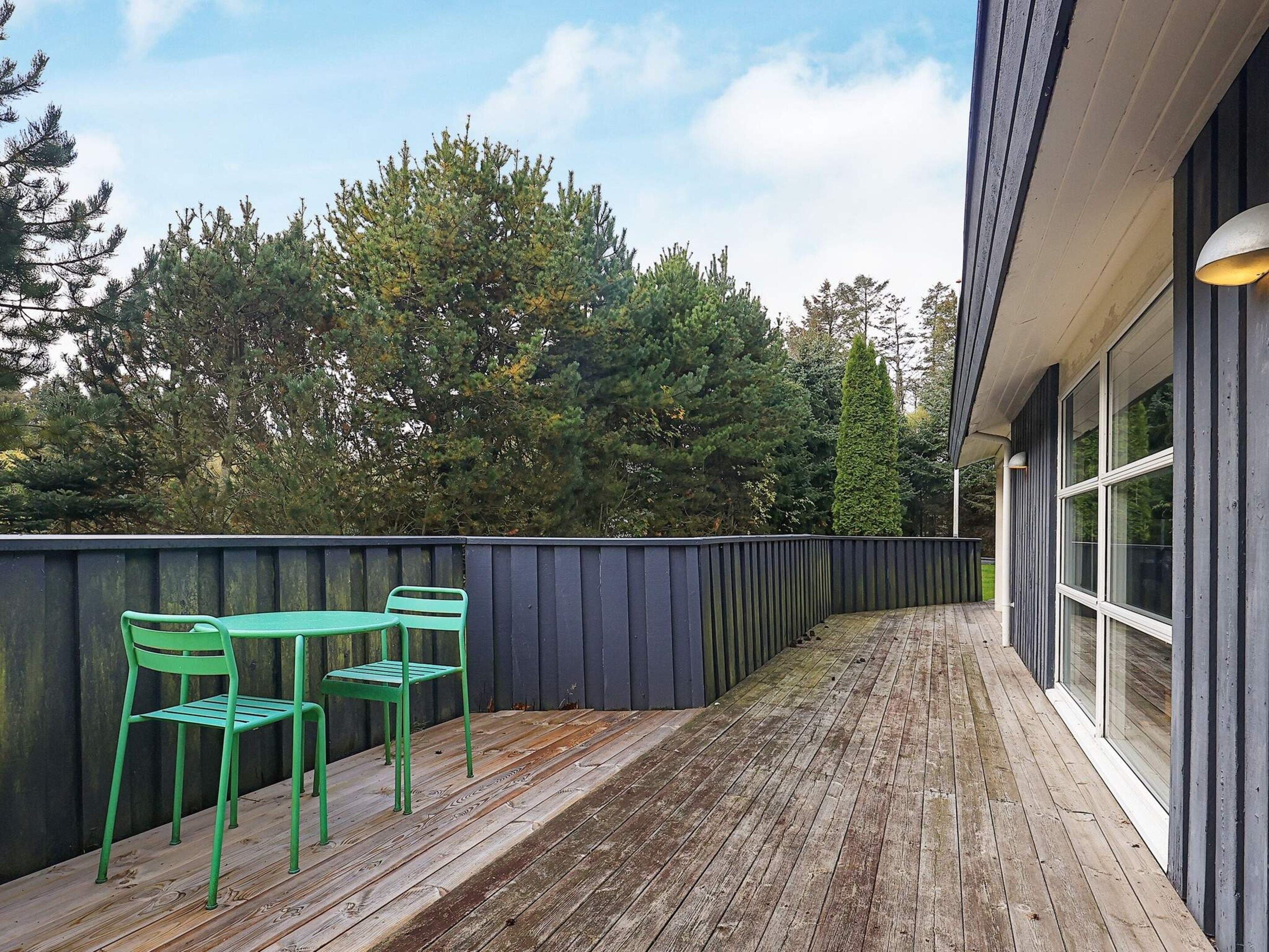 A wooden deck with two green chairs, a dark fence, and a building with large windows.